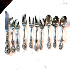 Könnte beinhalten: Eine Sammlung antiker Silberbestecke, darunter Gabeln, Löffel und ein Messer, auf einer weißen Marmoroberfläche angeordnet. Das Besteck hat verzierte florale Details an den Griffen und besteht aus glänzendem, silberfarbenem Metall.