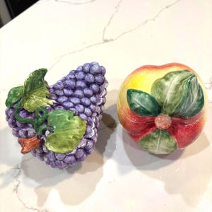 May include: Two ceramic fruit-shaped containers. One is a bunch of purple grapes with green leaves and a red stem. The other is a red, yellow, and green apple with green leaves and a pink bow.