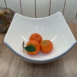 May include: A large, white, square-shaped ceramic bowl with curved, upward-sweeping sides. Inside the bowl are three bright orange tangerines, one of which features a small, attached green stem and a single green leaf, resting on the smooth surface.