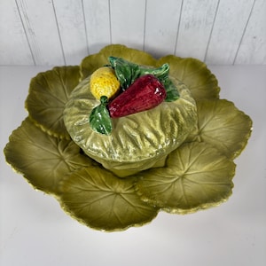 May include: A green ceramic serving dish shaped like a cabbage leaf with a smaller bowl in the center. The bowl is also shaped like a cabbage leaf and has a red pepper, a yellow lemon, and green leaves on top.