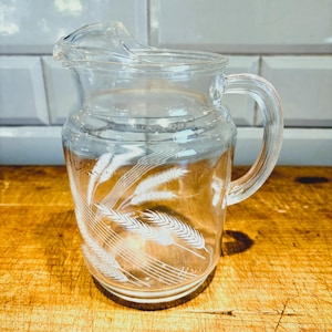 May include: Clear glass pitcher with a handle and a spout. The pitcher features a wheat stalk design etched on the front. The pitcher is sitting on a wooden surface.