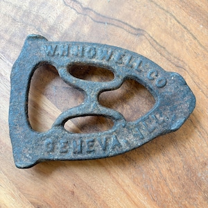 May include: A cast iron trivet with the words "Whitcomb Co. Geneva, N.Y." embossed on it. The trivet has a decorative design with a central opening and four smaller openings around it.