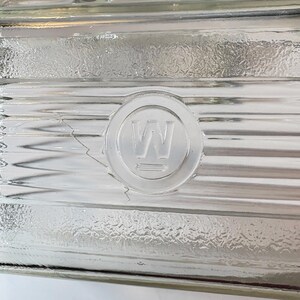 May include: Close-up of a clear glass block with a textured surface. A circular emblem with the letter "W" is embossed on the glass. The block has horizontal ridges and a slight crack.
