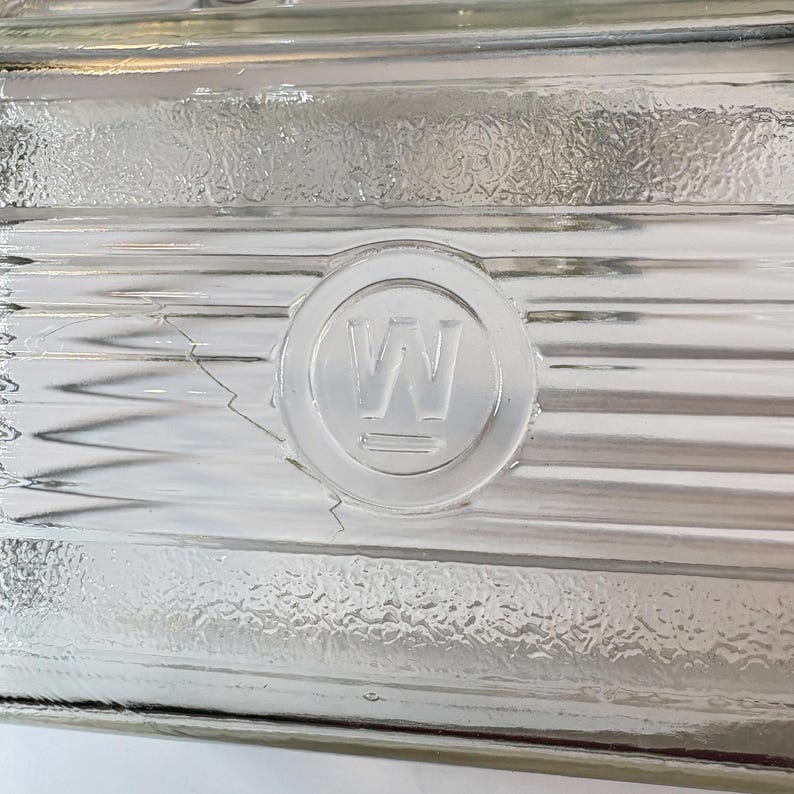May include: Close-up of a clear glass block with a textured surface. A circular emblem with the letter "W" is embossed on the glass. The block has horizontal ridges and a slight crack.
