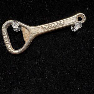 May include: A vintage silver bottle opener with the word "THORBEN" stamped on the handle. The opener has a loop at one end and a curved opening for removing bottle caps. The opener is on a black background.