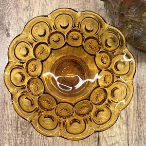 Op de afbeelding: Amberkleurige glazen schaal met een ster- en maanmotief. De schaal heeft een geschulpte rand en een centrale kom. Het transparante glas laat licht door. Het staat op een houten oppervlak.