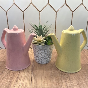 May include: Two ceramic watering cans, one pink and one yellow, with speckled finishes. They have a classic teapot shape with spouts and handles. A small gray pot with succulents sits between them. The background features a white tile pattern.