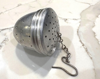May include: A silver tea infuser with a perforated, egg-shaped body and a hinged lid. A small chain and hook are attached for easy removal from a cup. The infuser is designed for steeping loose leaf tea.