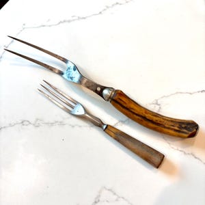 May include: Two antique serving forks. One fork has two long tines and a handle made of brown and tan material. The other fork has three tines and a wooden handle. Both forks are made of metal and are set against a white background.