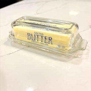 May include: A clear glass butter dish containing a rectangular stick of salted butter. The butter wrapper is yellow with blue text that reads Salted Butter, Sweet Cream, and Net Wt 4 oz (113 g). The dish has a removable lid with a handle.