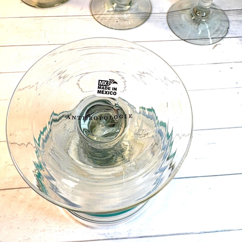 May include: Clear glass Anthropologie wine glasses with a wide bowl and a stem. The base of the glass has a teal accent. The bottom of the glass has a sticker that says "MX MADE IN MEXICO" and "ANTHROPOLOGIE".