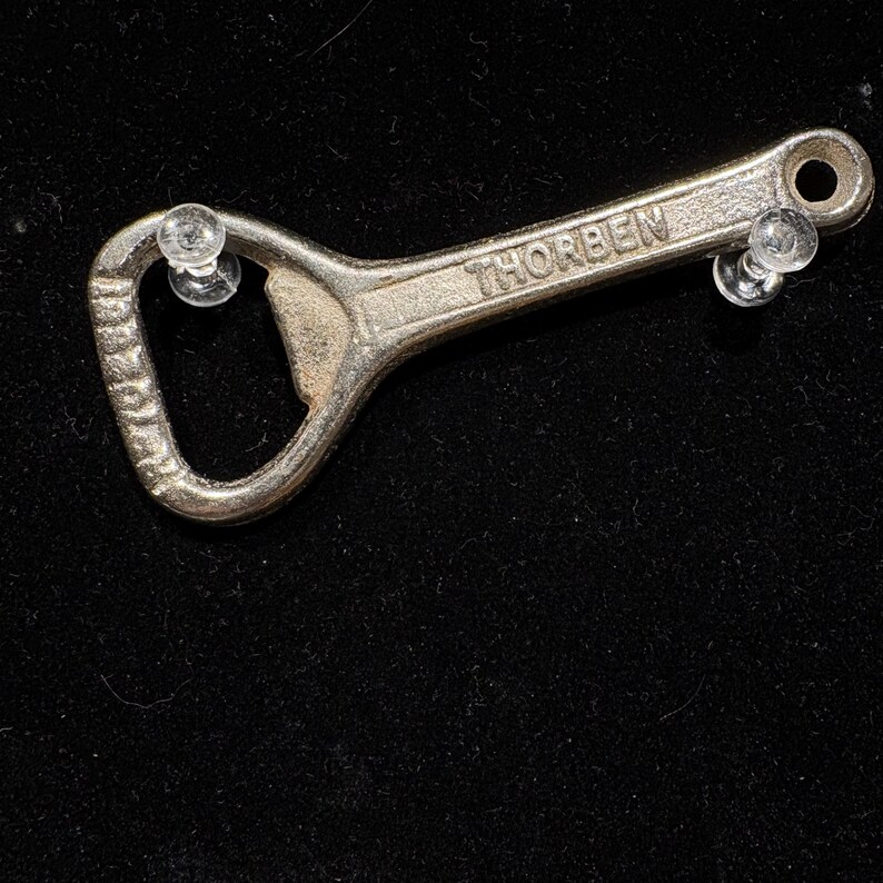 May include: A silver bottle opener with the name "THORBEN" engraved on the handle. The opener has a classic design with a loop at one end and a bottle cap remover at the other. The background is black.