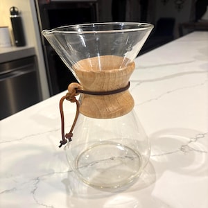 Może przedstawiać: Przezroczysty szklany ekspres do kawy z drewnianym kołnierzem i skórzanym paskiem. Karafka ma stożkową górę i zaokrąglone dno. Drewniany kołnierz jest jasnobrązowy. Skórzany pasek jest ciemnobrązowy.