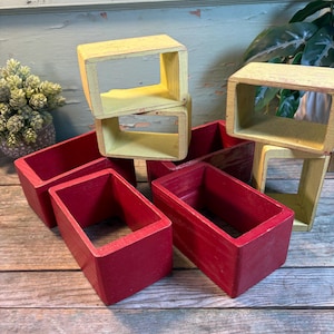 Puede incluir: Una colección de cajas rectangulares de madera en tonos rojos y amarillos. Las cajas están abiertas en ambos extremos, creando una forma rectangular hueca. Las cajas de madera están apiladas y dispuestas sobre una superficie de madera.