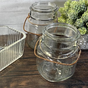 May include: Clear glass jars with wire handles and lids. The jars have a vintage aesthetic, with a slightly aged appearance. The jars are sitting on a wooden surface. The jars are suitable for storage or decorative purposes.