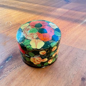 May include: A round, hand-painted trinket box with a floral design. The box is painted in shades of green, yellow, orange, and red.