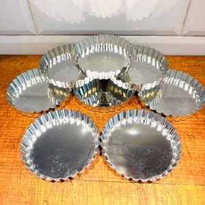 Puede incluir: Conjunto de siete moldes para tartas de metal de color plateado con bordes acanalados. Los moldes están apilados uno encima del otro sobre una superficie de madera.