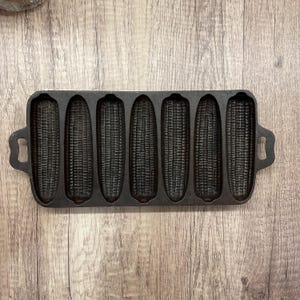 May include: A cast iron corn stick pan with six corn-shaped molds. The pan is black and has handles on each end. The molds are detailed to resemble corn on the cob. The pan is designed for baking corn bread.