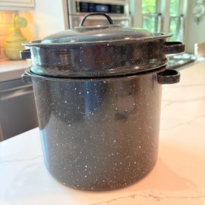 May include: A black speckled enamel stockpot with a matching lid and two side handles. The pot features a cylindrical body with a white speckled pattern throughout the dark surface, a rounded lid with a central handle, and two side loop handles.