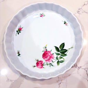 May include: White ceramic pie plate with a scalloped edge, decorated with pink roses and green leaves. The floral design is centered on the plate, with additional rose buds scattered around the rim. The plate is round.