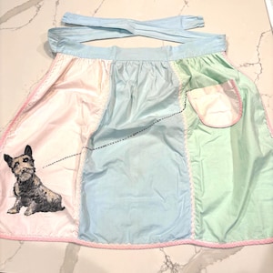 May include: A vintage-style apron with a patchwork design in pastel pink, blue, and green. The apron features a charming embroidered dog on the pink panel, a pocket, and pink trim. The apron has light blue ties.