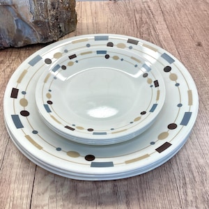 May include: A stack of white ceramic plates and bowls with a geometric pattern of brown, tan, and blue shapes. The plates are arranged on a wooden surface, with a piece of stone in the background.