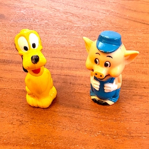 Puede incluir: Un perro de juguete de plástico amarillo con la lengua roja y un cerdo de juguete de plástico azul con un sombrero azul y un mono.