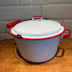 May include: White enamelware pot with a lid and red accents. The pot has a red handle on the lid and red handles on the sides. The pot sits on a wooden surface. The pot is a vintage kitchenware item.