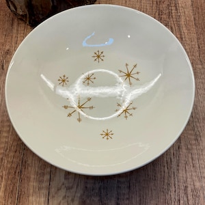 Könnte beinhalten: Eine runde, cremeweiße Keramikschale mit einem verstreuten Muster aus goldfarbenen Sternen- oder Schneeflockenmotiven auf der Innenseite. Die Schale hat eine glatte, glänzende Oberfläche und eine einfache, kreisförmige Form mit breitem Rand.