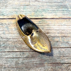 May include: A gold-colored ashtray in the shape of a shoe, with a detailed leaf design. The ashtray has a dark interior and is set against a weathered wooden surface. The shoe-shaped ashtray is a decorative item.