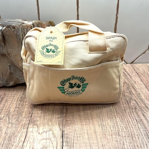May include: A cream-colored canvas bag featuring a green Cabbage Patch Kids Airways logo on the front. A matching tag is attached to the handle, displaying the text SPAIN VIA Cabbage Patch Kids AIRWAYS with a graphic of a stork carrying a bundle.