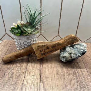 May include: A weathered wooden tool, marked with "OTTO BERNZ CO ROCHESTER", rests on a wooden surface. A small potted succulent and a cluster of pale blue crystals are also visible. The tool features a handle and a rectangular head.