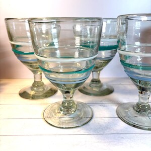 May include: Set of four clear glass goblets with a wide bowl and a stem. Each goblet is adorned with horizontal bands of white, teal, and light blue. The glasses are arranged on a white surface, showcasing their design.
