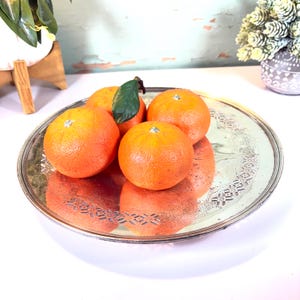 Puede incluir: Una bandeja plateada ornamentada exhibe cinco naranjas de color naranja brillante. Una naranja tiene una hoja verde. La bandeja refleja las naranjas. El fondo es una pared azul claro y una superficie blanca.