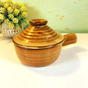 May include: A handmade, speckled ceramic casserole dish with a lid and handle. The dish is a warm brown color with darker brown accents. The lid has a rounded shape with concentric ridges. The handle is a solid, cylindrical shape. A decorative vase with greenery is in the background.