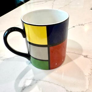 May include: A ceramic mug with a black handle and a white interior. The exterior is divided into colorful squares of yellow, navy blue, gray, red, and green, separated by black lines. The mug sits on a white countertop.