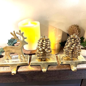 Puede incluir: Tres sujetadores dorados para medias navideñas. Uno es un reno y los otros dos son piñas. Cada soporte tiene una base cuadrada y un gancho para colgar las medias. El fondo incluye velas encendidas.