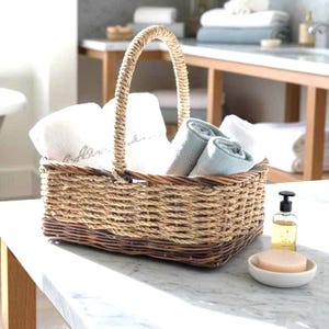 May include: A rectangular woven wicker basket with a tall arched handle, containing several folded white and light blue towels. The basket features a natural tan upper section and a darker brown base, providing a sturdy storage solution.