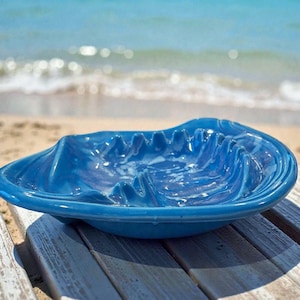 May include: A vibrant blue ceramic ashtray with a wave-like design. The ashtray is placed on a wooden surface with a beach and ocean in the background. The ashtray's interior has a marbled effect.