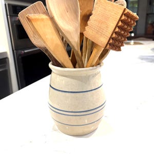 Könnte beinhalten: Eine Sammlung von Küchenutensilien aus Holz, darunter Löffel und Spatel, in einem cremefarbenen Keramiktopf mit blauen Streifen. Die Utensilien sind in verschiedenen hellbraunen Farbtönen gehalten.