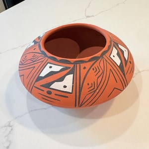 May include: A low, rounded terracotta bowl with a wide opening. The exterior surface features a matte finish in a reddish-orange hue, decorated with intricate geometric patterns in black and white, including triangles, lines, and small dots.