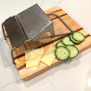 May include: A stainless steel cheese slicer with a wooden cutting board. Sliced cheese and cucumber are arranged on the board. The board has a light wood grain with dark wood stripes.