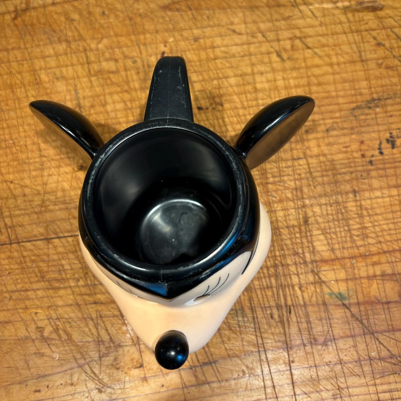May include: A black and beige ceramic mug shaped like a cartoon character's face. The mug has a black handle and black ears extending from the sides. The character's nose is a black sphere, and the face is beige with black details.