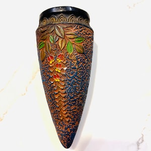 May include: A decorative, cone-shaped vase with a textured brown surface and blue accents. The vase features a floral design with green leaves and red and yellow flowers. The top rim is black.