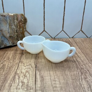 Puede incluir: Dos recipientes de vidrio lechoso blanco, uno es un cuenco pequeño con asa lateral y el otro una jarra para crema con pico vertedor y asa lateral. Ambas piezas presentan un acabado liso y opaco con una forma redondeada para servir.