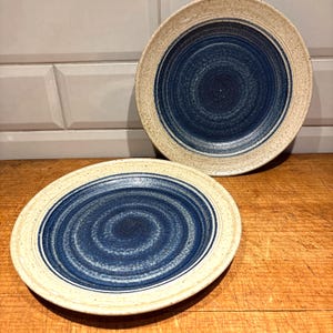May include: Two ceramic plates with a spiral design. The plates have a cream-coloured rim and a deep blue centre with a swirling pattern. The plates are on a wooden surface.