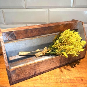 Puede incluir: Una caja de herramientas de madera rústica con asa, llena de un ramo de flores amarillas brillantes atadas con hilo. La caja de herramientas tiene un aspecto envejecido y desgastado, con vetas de madera visibles y un interior azul claro.