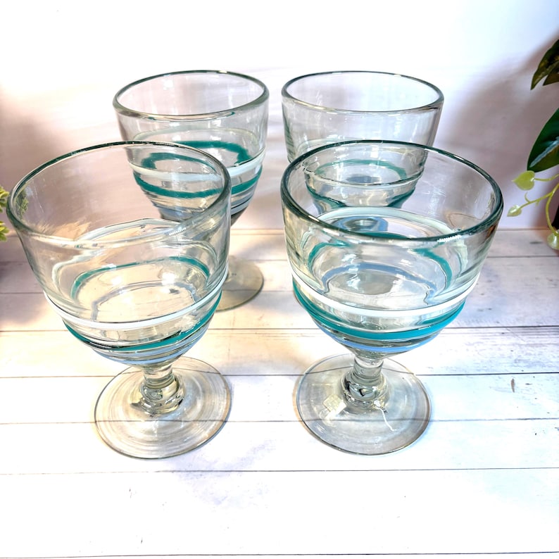 May include: Four clear glass goblets with a teal, white, and light blue spiral design around the bowl. The goblets have a stemmed base and are arranged on a white wooden surface. The glassware is suitable for serving beverages.