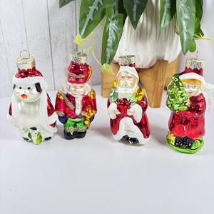 Puede incluir: Cuatro adornos navideños de vidrio vintage que representan un pingüino, un soldado, Papá Noel y una niña que sostiene un árbol de Navidad. Los adornos son rojos, blancos, verdes y azules.