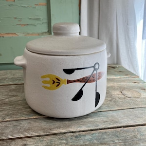 May include: A speckled, off-white ceramic canister with a lid and two small handles. The canister features a yellow fork, brown spoon, and black spoon design. The canister is sitting on a wooden surface.